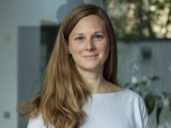 Das Foto zeigt ein junges, vermutlich weibliches menschliches Individuum. Das volle lange Haar ist zur rechten Seite nach vorn gekämmt und verdeckt an dieser Stelle den hellblauen Pullover, der trefflich zu den blauen Augen passt. Das Individuum steht vor einem schemenhaft verzogenen Hintergrund, der die Örtlichkeit nicht klar zu erkennen gibt.