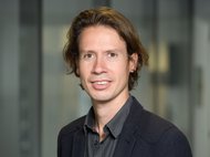 Person mit welligem braunem Haar, gekleidet in ein dunkles Hemd und einen Anzug, steht vor einer Glaswand im Büro.