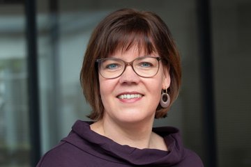 Eine Frau mit mittellangen braunen Haaren, bebrillt, trägt lila Oberteil und Ohrringe, steht vor einem unscharfen Hintergrund.