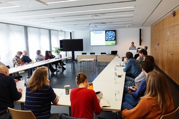 In einem hellen Konferenzraum verfolgen mehrere Personen einen Vortrag über Gleichheit, Vielfalt und Inklusion auf einem Bildschirm.