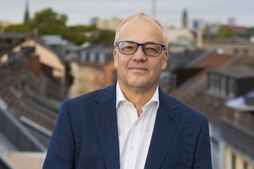 Mann mit Brille im Anzug mit offenem Hemd vor dunkliem Hintergrund stellt sich dem Fotografen.