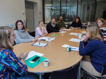 Regelmäßig finden Besprechungsrunden in den Servicegruppen statt – hier die Verwaltung bei ihrem wöchentlichen Meeting. Gruppenbesprechung an einem ovalen Tisch, Dokumente und Getränke sind verteilt. Fenster im Hintergrund.
