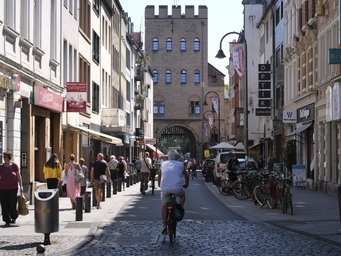 Die Severinstraße in der Nähe des Instituts bietet Einkaufs- und Ausgehmöglichkeiten In der belebten Einkaufsstraße fahren Radfahrer und gehen Fußgänger, flankiert von Geschäften und einem historischen Torbogen im Hintergrund.
