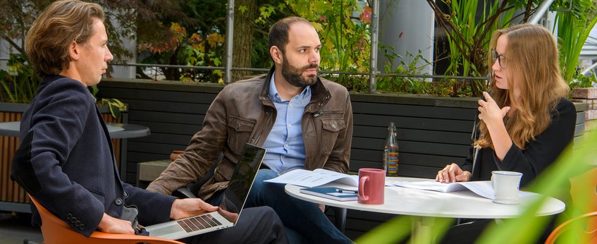 Drei Personen sitzen an einem runden Tisch im Freien und führen ein Gespräch. Auf dem Tisch befinden sich ein Laptop, Dokumente, eine Flasche und eine Tasse. Im Hintergrund sind Pflanzen zu sehen.