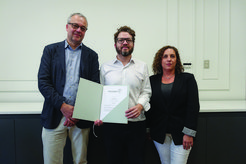 Three people pose indoors, the person in the middle displays a certificate from the Max Planck Institute, suggesting an award or achievement.