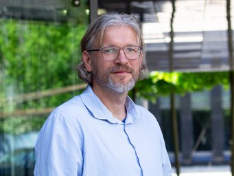 A person with medium-length hair wearing a light blue shirt stands in front of a modern building with a glass facade and green trees in the background.
