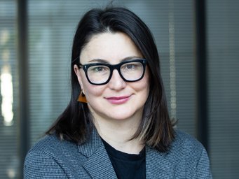 Person with dark, shoulder-length hair, dressed in a checkered blazer and black shirt, positioned before closed venetian blinds.