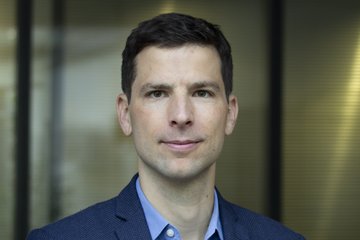 A person is depicted standing in front of a glass wall with a blurred background. The person is wearing a structured blue jacket and light blue shirt.