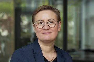 Person wearing a blue blazer and black shirt, wearing a gold chain. The background shows a glass wall with blurred green and white details.