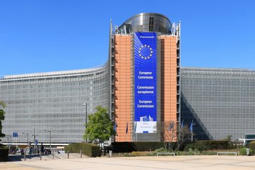The European Commission headquarters in Brussels, showcasing a large banner with the EU emblem and the commission's name in English, French, and German. The building has a modern architectural design with glass and steel elements, surrounded by greenery and a clear blue sky.