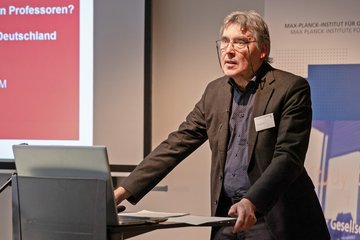 Ein Mann steht auf einem Podium und hält eine Präsentation, während im Hintergrund Folien zu sehen sind.