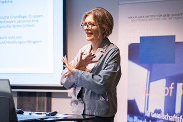 Eine Frau mit kurzen Haaren, Brille und hellgrauem Blazer spricht an einem Rednerpult. Sie gestikuliert mit den Händen, hinter ihr befindet sich eine Präsentationsleinwand, auf der ein Text in deutscher Sprache angezeigt wird.