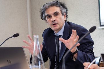 Ein Mann im Anzug gestikuliert, während er an einem Konferenztisch spricht, vor ihm stehen ein Laptop und eine Glasflasche mit Wasser.