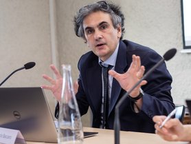 A man in a suit gestures while speaking at a table with a laptop and a water bottle.