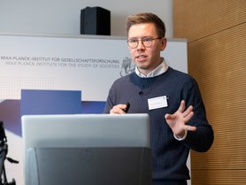 A man is presenting at a conference, holding a remote control, with a laptop in front of him. He is wearing glasses and a sweater, and there is a sign in the background referencing the Max Planck Institute for the Study of Societies.