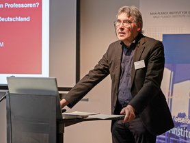 A speaker presenting at a conference or seminar, standing behind a laptop with a slide presentation in the background.