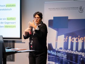 A woman is presenting, gesturing with her hands, in front of a projector screen displaying text in German. A banner for the Max Planck Institute for the Study of Societies is in the background.