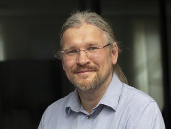 An individual with long gray hair tied back, wearing a light blue, buttoned shirt, positioned in an indoor setting with soft lighting.