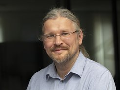 An individual with long gray hair tied back, wearing a light blue, buttoned shirt, positioned in an indoor setting with soft lighting.