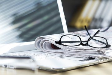 Auf einem aufgeklapptem Laptop liegt eine gefaltete Zeitung, auf der Zeitung liegt eine Lesebrille.