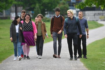 A group of seven people is walking along a wide gravel path flanked by green meadows and scattered trees. They are dressed casually, and some of them are carrying backpacks.