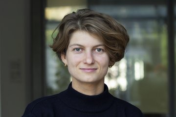 Woman with light brown short hair, wearing a dark sweater and gold earrings. Background slightly blurred with glass texture.
