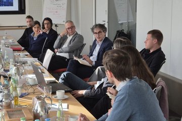 A group of professionals sit around a conference table in discussion. Laptops, notebooks, and drinks are on the table, indicating a collaborative meeting.