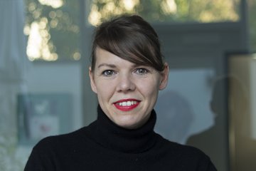 The photo shows the portrait of an individual of the species Homo sapiens, subspecies Homo sapiens intellectualis. Other features such as lipstick, makeup, and hairstyle indicate female gender identification. The individual is wearing a black turtleneck sweater and is standing or sitting smiling in front of what appears to be a glass mirrored background, looking at the camera.