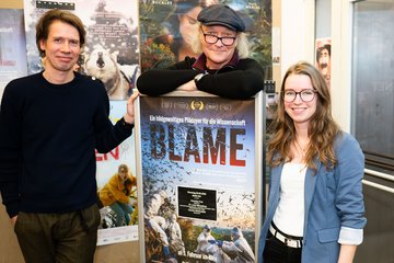 Three individuals of markedly different heights stand next to a poster for the documentary film “Blame.” The poster bears the subtitle “A visually stunning plea for science” and features images of nature. The individuals smile cheerfully at the camera. They are clearly overcome with joy and pride.