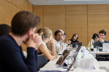 The image shows a group of individuals of the species Homo sapiens intellectualis sitting at simple tables in a modern-looking space, clearly engrossed in a lively discussion. They all have a so-called laptop placed on the table; the purpose is likely to prepare arguments during the discussion or to present texts they have already prepared.
