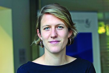 The photo shows a member of the species Homo sapiens intellectualis with short blond hair and hoop earrings, wearing a dark blue sweater, against a blurred background in an interior setting featuring a window and a poster.