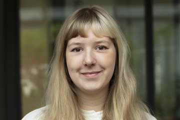 A young specimen of the species Homo sapiens intellectualis, with long blond hair falling forward over her chest, wearing a light-colored, collarless sweater, smiling in a friendly yet hesitant manner, standing or sitting against a blurred window background, looks directly at the viewer.
