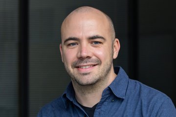 A male individual of the species Homo sapiens intellectualis, smiling warmly at the viewer, with thinning hair that is barely there and the hint of sprouting facial hair, wearing a blue shirt with an open collar, stands or sits in front of an indefinably blurred background that could possibly be interpreted as a curtain.