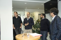Five men in casual clothing converse in an office setting, standing around a table with glasses and documents, surrounded by plants and artwork.