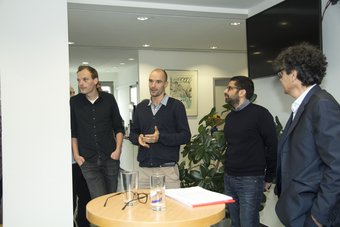 New researchers at the MPIfG Five men in casual clothing converse in an office setting, standing around a table with glasses and documents, surrounded by plants and artwork.