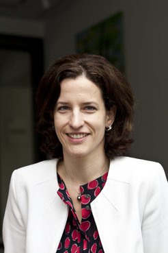 A woman dressed in a white blazer and a red-black patterned blouse, featuring a pearl pendant necklace and small hoop earrings, stands indoors.