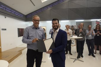 Felipe González and Jens Beckert In a contemporary hall, two men in business attire present a certificate, while several attendees stand in the background, applauding the achievement.