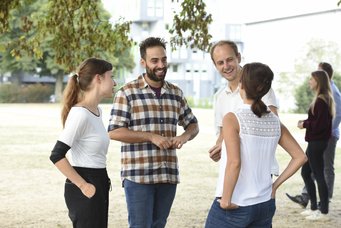 Four people engaged in conversation outside, surrounded by greenery, with buildings visible in the background.