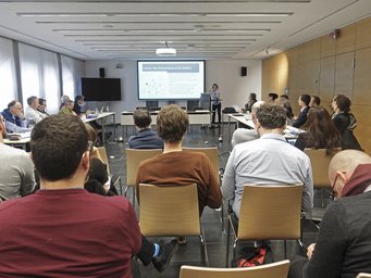 Conference on the politics of growth and stagnation The photo provides a glimpse into a conference room where members of the species Homo sapiens intellectualis are seated at rectangular tables, following the event. In the background, an oversized monitor can be seen, presumably used for presentation purposes by the person in charge.