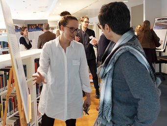 Members of the Scientific Advisory Board and doctoral students discuss posters presented during an MPIfG evaluation. In a library setting, two individuals interact near informational displays, while other attendees browse and converse in the background.