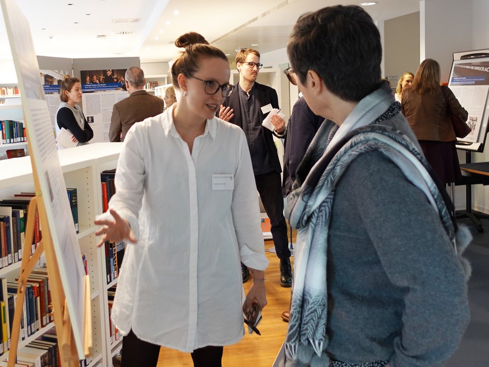 In a library setting, two individuals interact near informational displays, while other attendees browse and converse in the background.