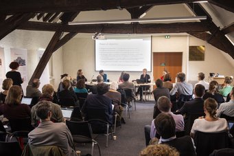 Tenth-anniversary and closing conference of Maxpo in October 2022 in Paris Conference participants listen attentively to a presentation. Panel members discuss at a table in the foreground. A screen with text is visible in the background. Some audience members work on laptops. The scene takes place in a room with wooden beams.