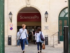 Entrance to SciencesPo, a renowned university, with several people entering or leaving the campus. Signs and lanterns are visible on the wall in the foreground.