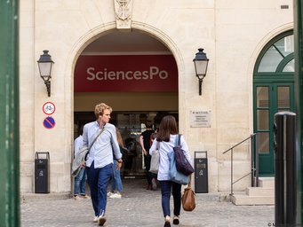 Foto von Marta Nascimento, Sciences Po (2019), Eingang Gebäude Rue de la Chaise Entrance to SciencesPo, a renowned university, with several people entering or leaving the campus. Signs and lanterns are visible on the wall in the foreground.