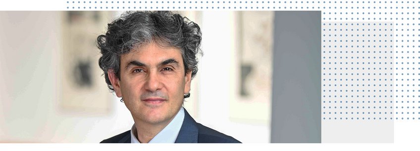 Prof. Dr. Lucio Baccaro Individual with curly hair, dressed in a dark suit against an abstract backdrop.