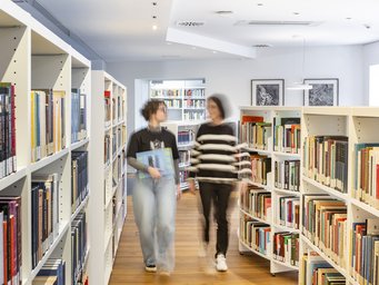 Two people walking along an aisle in a modern library, surrounded by white bookshelves filled with various books, with wooden flooring and framed pictures on the wall.