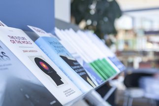 Eine Reihe von Büchern, die in einem Regal präsentiert werden. Die Titel umfassen "Uncertain Futures" und "The Future of the World". Im Hintergrund ist eine unscharfe Bibliotheksumgebung mit einer Person zu sehen.