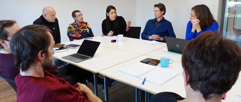 Regelmäßig trifft sich der geschäftsführende Direktor mit den Vertreterinnen und Vertretern der Servicegruppen, der Ausschüsse, des Betriebsrats und der Forschungskoordination im Managementausschuss. In einem hellen Raum sitzen Personen an einem Tisch und tauschen aktiv Ideen aus. Um sie herum liegen Laptops, Notizbücher und Kaffeetassen verstreut, was auf eine gemeinsame Arbeitssitzung hindeutet.