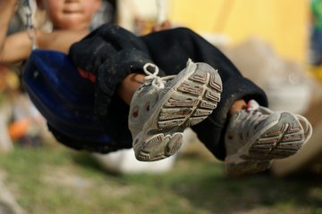 Close-up of a child's feet in white sneakers, sitting on a blue swing, with blurred outdoor background.