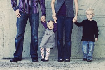 A group of four, comprising two adults and two children, stand side by side against a concrete backdrop. The adults, dressed in casual attire, hold the children's hands, conveying a sense of togetherness and familial bond.